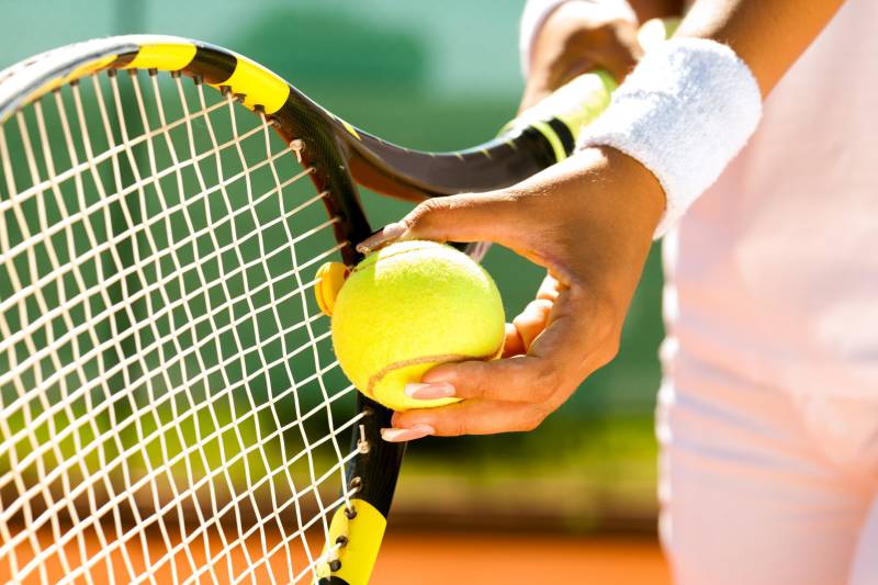 A tennis player grips a bright yellow ball against the strings of their racket on a sunny court.
