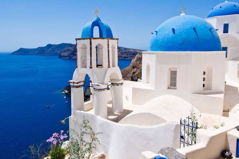 Iconic blue-domed churches perch above the Aegean Sea on Santorini, Greece, under a brilliant blue sky.