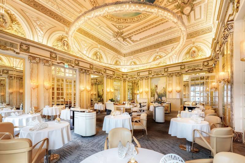 Elegant dining tables are set with white linens and ornate chairs in a grand, gilded room illuminated by a massive circular chandelier.