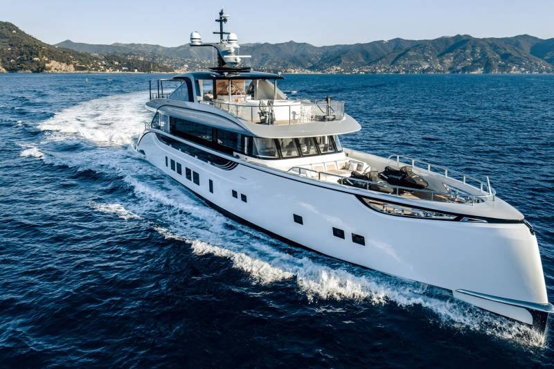 A sleek white yacht glides through the deep blue sea, leaving a foamy wake against a backdrop of lush green hills.