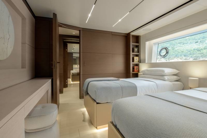 Luxurious twin beds with soft grey throws are bathed in natural light from a large porthole window overlooking a vibrant underwater landscape.