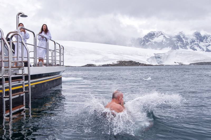 A man plunges into frigid Antarctic waters from a ship, while others in robes watch from the deck.