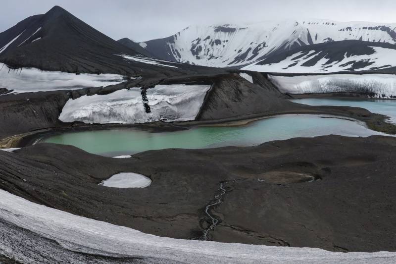 A stark landscape of dark volcanic rock and snow-capped mountains surrounds a serene, turquoise lake.