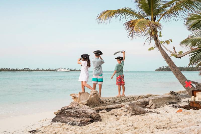 Three children dressed as pirates stand on a sandy beach, looking out at the turquoise ocean with a treasure chest nearby.