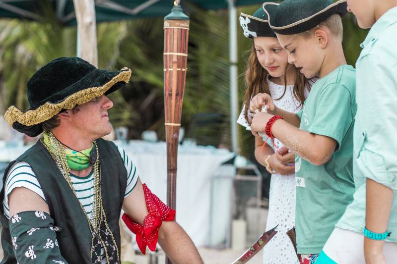 The image shows a group of people dressed in traditional cultural attire, with one individual wearing a distinctive hat and holding a ceremonial staff, while the others appear to be observing or participating in some kind of cultural event or celebration, set against a backdrop of palm trees and other tropical vegetation.