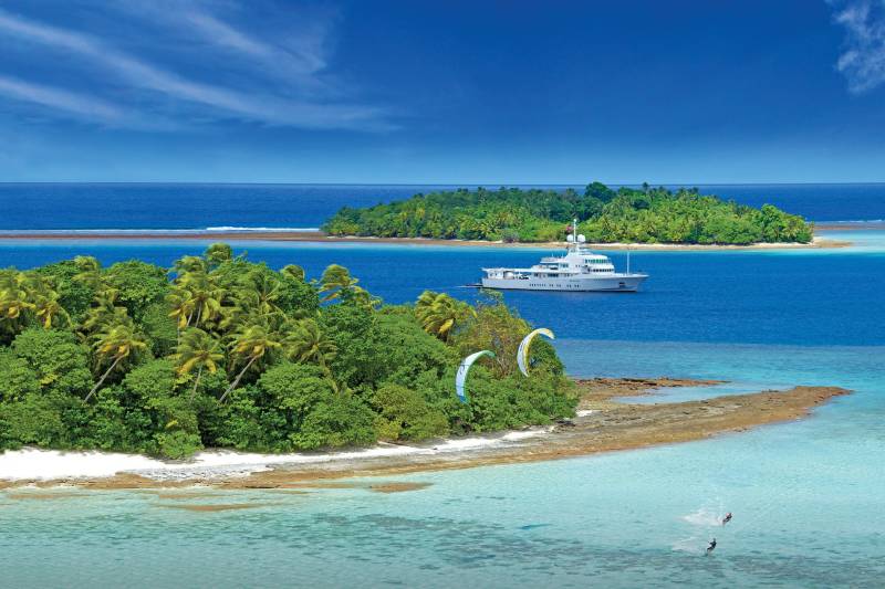 Kiteboarders enjoy the crystal-clear waters surrounding a lush tropical island with a luxury yacht anchored offshore.