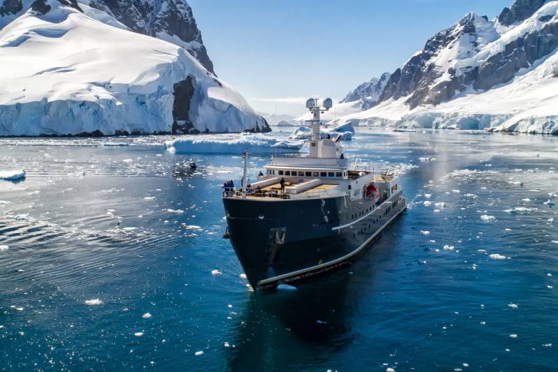 A majestic yacht navigates through icy Antarctic waters, dwarfed by towering snow-capped mountains.
