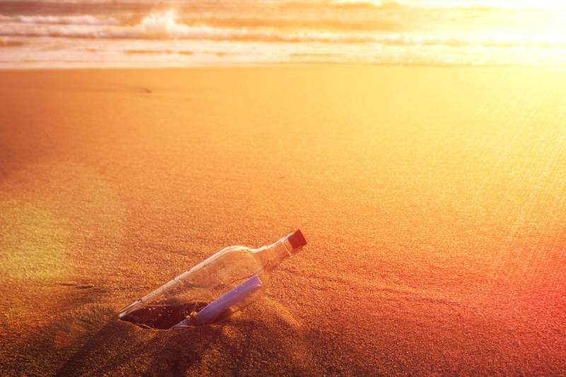 A message in a bottle rests on a sandy beach as the sun sets over the ocean.