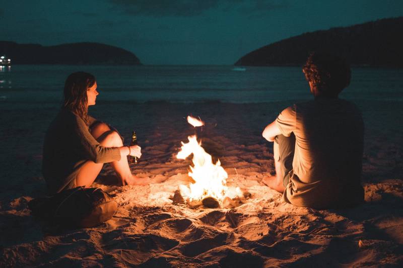 Two figures sit by a crackling bonfire on a sandy beach as the twilight sky meets the ocean.