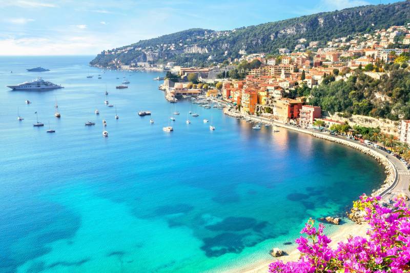 Luxury yachts and sailboats dot the azure waters of a stunning Mediterranean bay lined with colorful coastal villages.