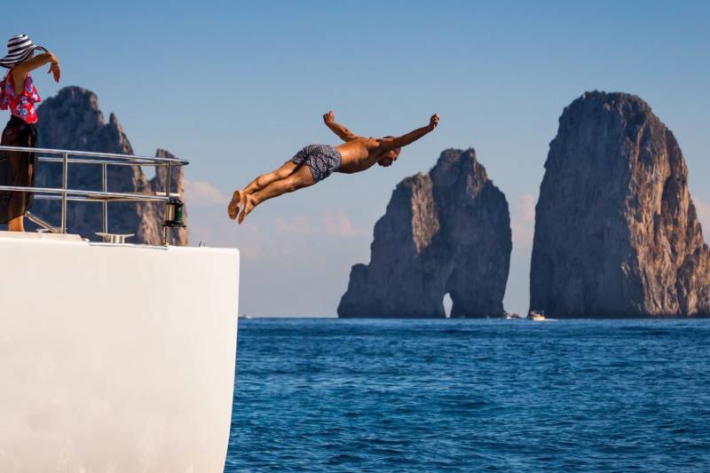 A man dives from a yacht into the azure waters near the iconic Faraglioni rock formations.