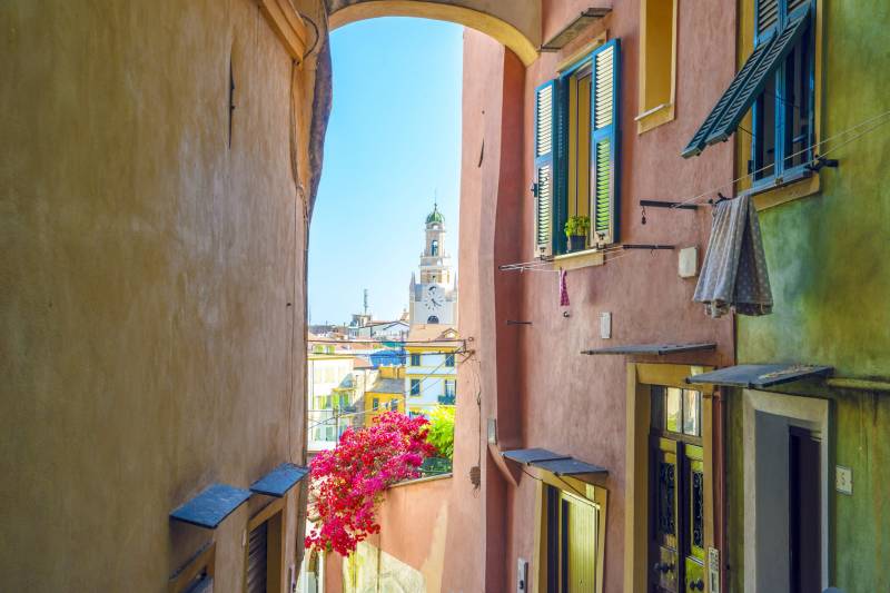 A narrow alleyway frames a view of colorful buildings and a distant clock tower under a bright blue sky.