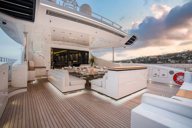 Luxurious yacht deck dining area with plush seating and elegant table setting overlooks a picturesque coastal town at sunset.