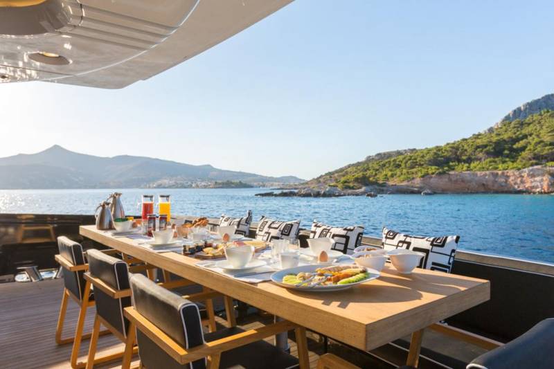 A beautifully set table awaits a delightful breakfast on a yacht overlooking a serene Mediterranean coastline.
