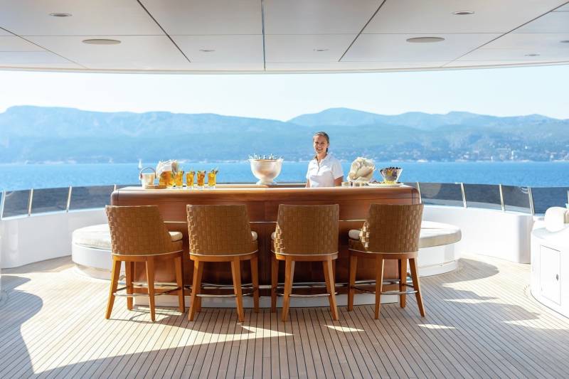 A smiling bartender serves refreshing drinks at a luxurious yacht bar overlooking a stunning coastal landscape.