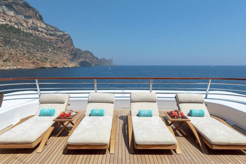 Luxurious chaise lounges await on a yacht deck, offering a serene view of a rugged coastline and the vast blue ocean.