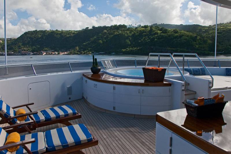 Relax on the deck of this luxurious yacht, complete with a soothing hot tub and comfortable lounge chairs, overlooking a lush green coastline.