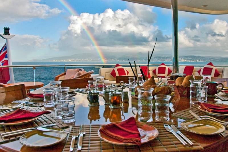 A rainbow arches over a beautifully set dining table on a yacht, overlooking a coastal landscape.