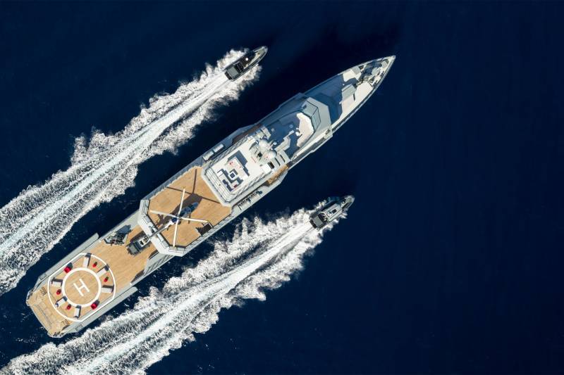 A sleek superyacht cruises through deep blue waters, accompanied by two agile speedboats leaving white wakes.
