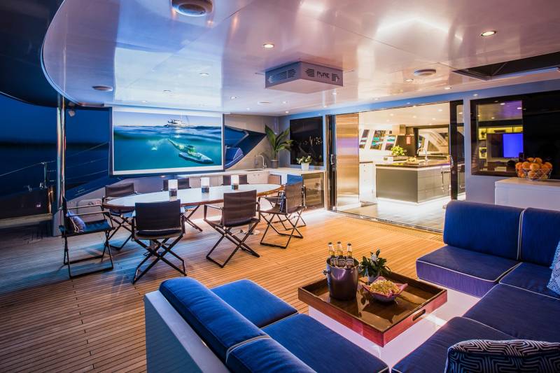 This luxurious yacht deck offers an open-air cinema experience with a large screen, comfortable seating, and a fully equipped galley visible through sliding doors.
