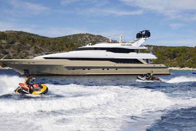 A luxury yacht anchors offshore as two jet skiers enjoy the crystal-clear waters of a Mediterranean island.
