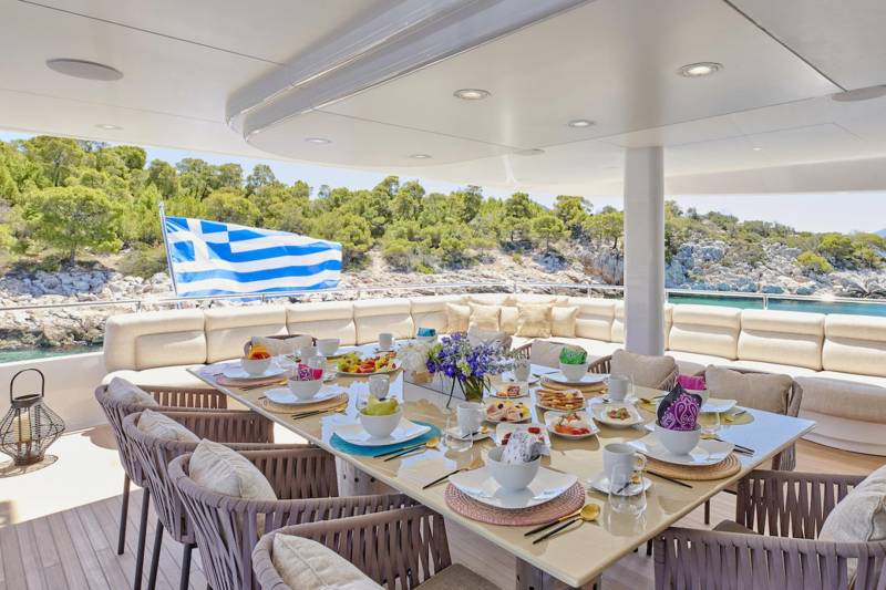 A beautifully set dining table on a yacht deck overlooks a picturesque Greek island coastline.