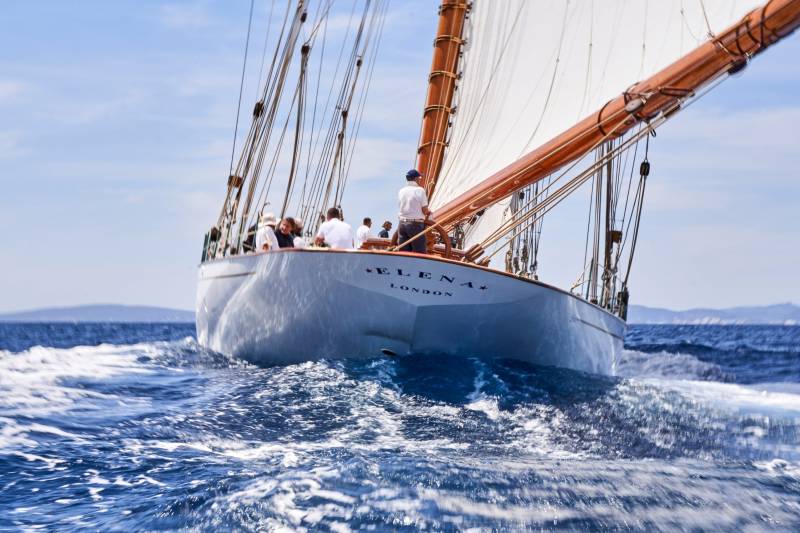 A majestic schooner named Elena cuts through the deep blue sea, its sails billowing under a clear sky.
