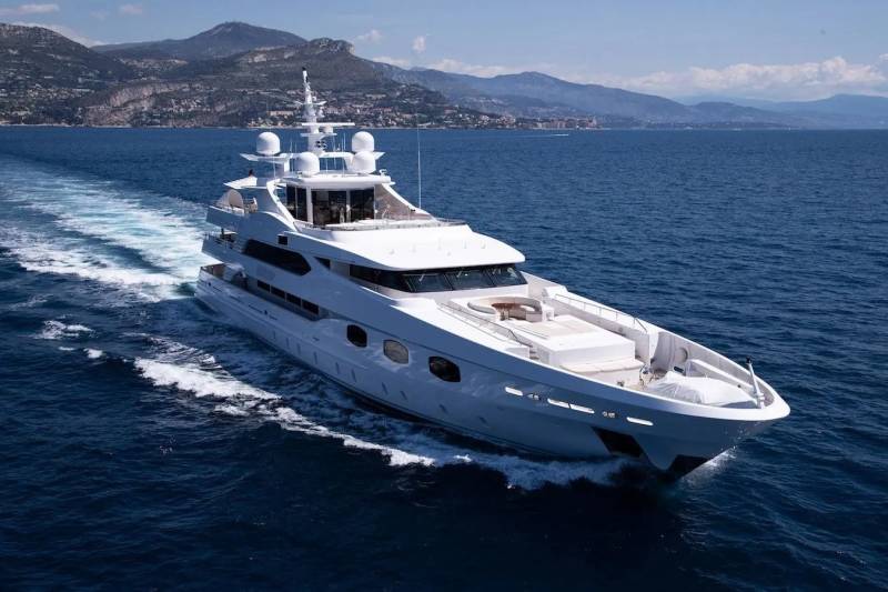 A sleek white yacht glides through the deep blue ocean, leaving a foamy wake behind it as mountains line the distant coast.