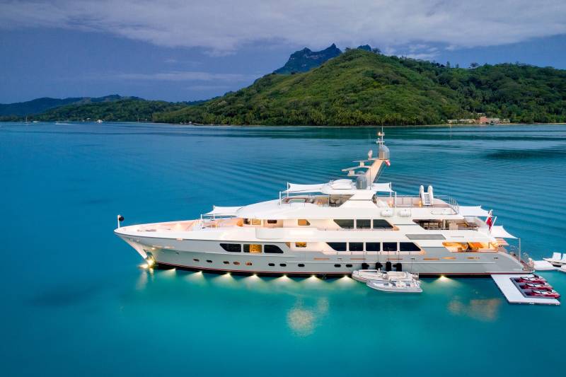 A luxurious white yacht anchors in the crystal-clear turquoise waters offshore a lush, mountainous island.