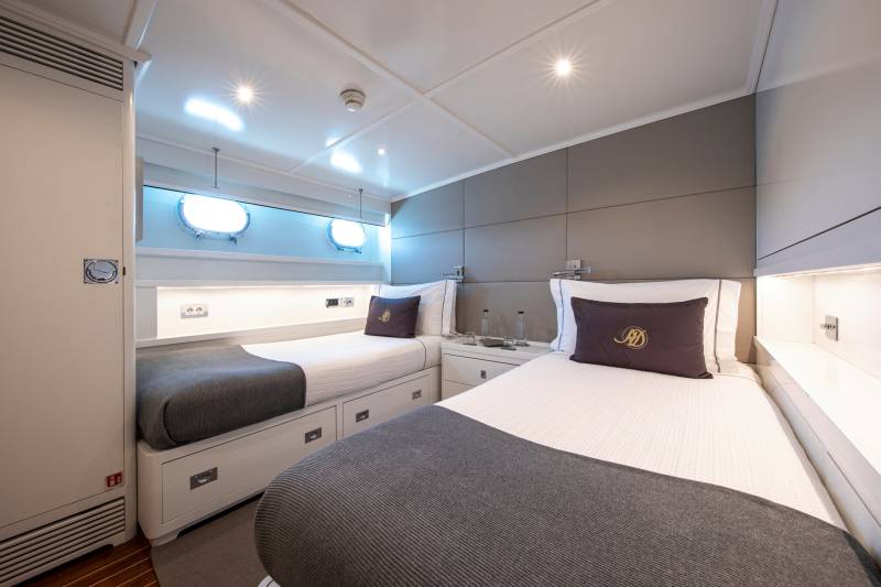Two twin beds with dark grey throws and decorative pillows are neatly made in this modern yacht cabin.