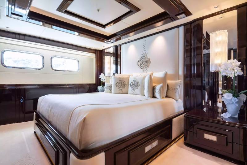 Luxurious yacht bedroom features a plush king-size bed, elegant rose-embroidered pillows, and rich dark wood accents.