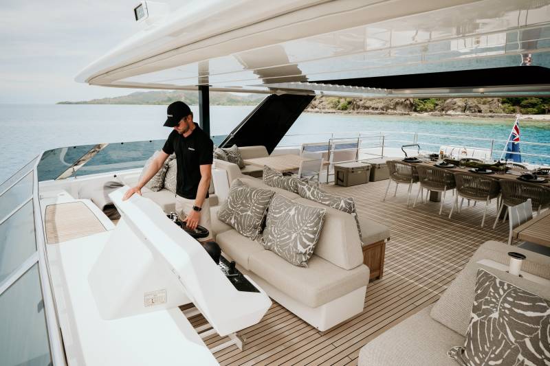 A man in a cap steers a luxury yacht through calm turquoise waters near a lush, tropical island.