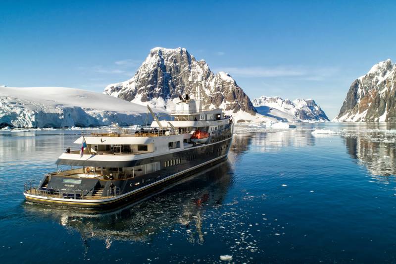 A sleek expedition ship navigates icy Antarctic waters beneath towering snow-capped mountains.