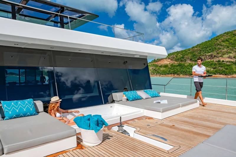A woman relaxes in a jacuzzi on a yacht's deck while a server brings drinks, with a lush green island in the background.