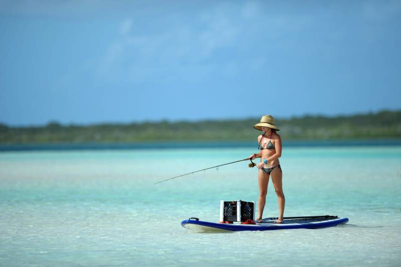 A woman in a bikini and straw hat fishes from a paddleboard in shallow, clear turquoise water.