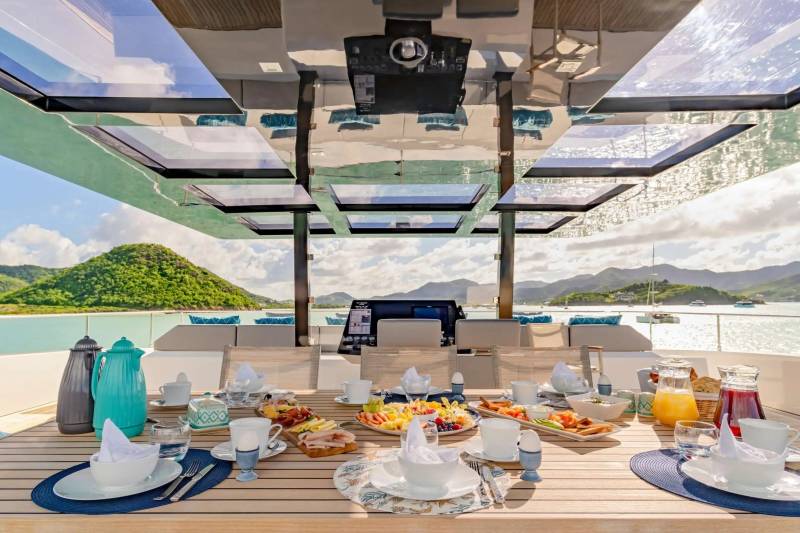 A luxurious breakfast spread awaits on a yacht deck, overlooking a serene tropical bay.