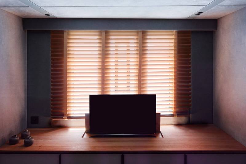 A minimalist room features a sleek television set on a wooden platform beneath slatted blinds casting warm light.