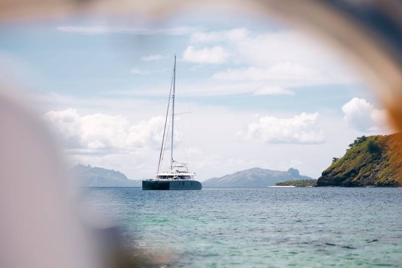 A sleek catamaran glides across turquoise waters near a lush, mountainous island under a cloudy sky.