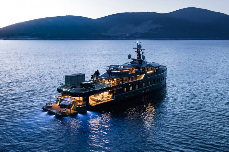A luxurious yacht with illuminated interiors and a rear deck pool rests on calm, dark blue waters under a twilight sky.