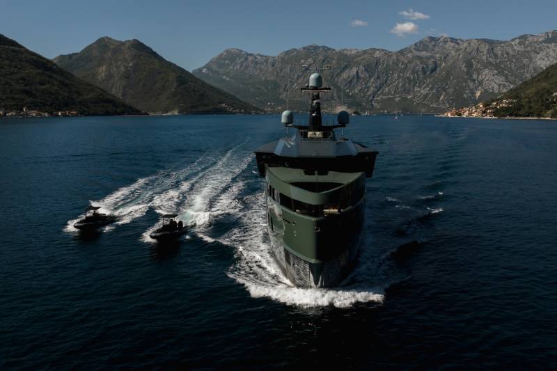 A sleek, dark green yacht cuts through the deep blue water, flanked by smaller escort boats, with a picturesque mountain village nestled on the shore.