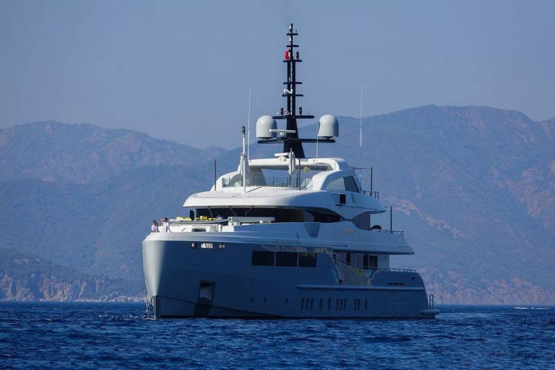 A sleek superyacht cruises through calm blue waters against a backdrop of hazy mountains under a clear sky.