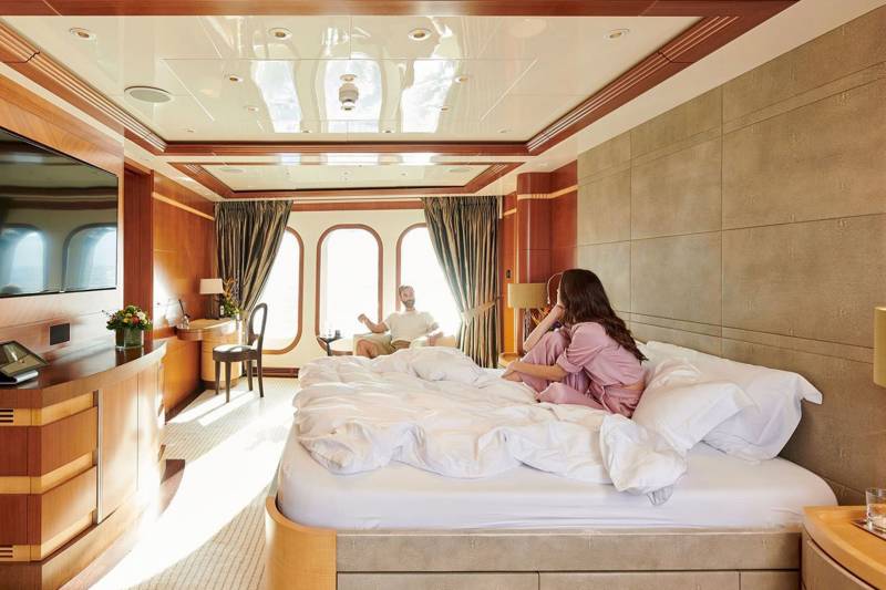 A couple relaxes in a luxurious yacht cabin with large arched windows overlooking the sea.