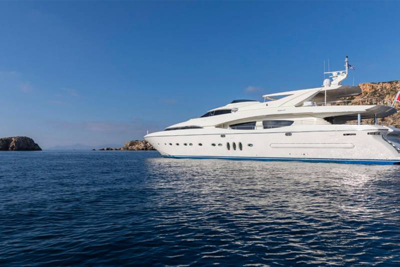A luxurious white yacht glides through the deep blue waters near a rocky Mediterranean coastline under a clear sky.