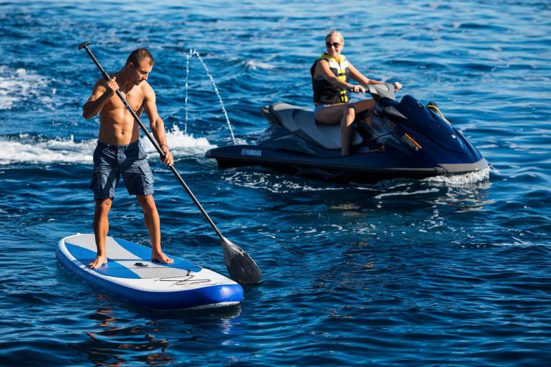 A man paddleboards on a calm sea while a woman rides a jet ski in the background.