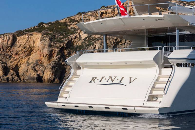 A luxurious yacht with the name "R.I.N.I.V." emblazoned on its stern cruises past a rocky, sun-drenched coastline.