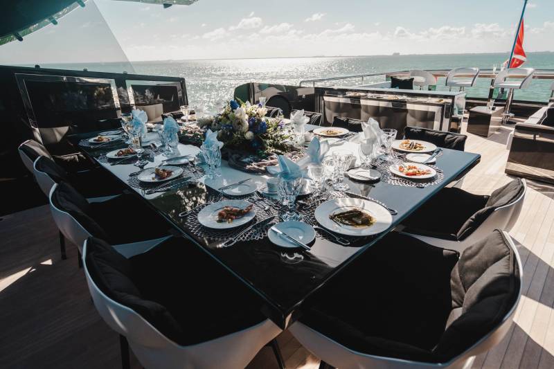 A beautifully set dining table on a yacht overlooks a sparkling ocean under a bright, cloudy sky.