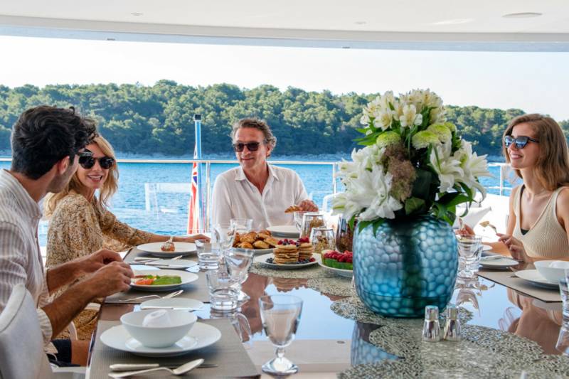 A group enjoys a luxurious breakfast spread on the deck of a yacht, surrounded by the sparkling blue sea and lush green islands.