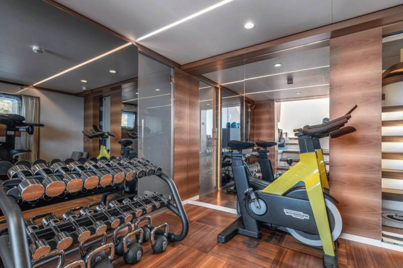 A modern yacht gym features a gleaming dumbbell rack and sleek stationary bikes set against rich wood paneling.