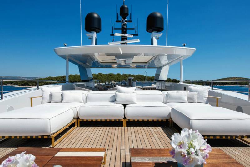 Luxurious white lounge seating and wooden tables adorn the deck of a yacht under a clear blue sky.
