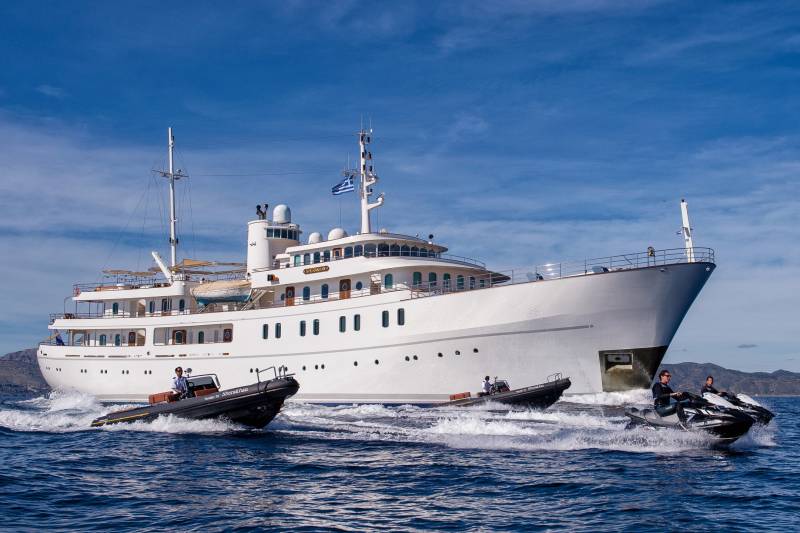 A sleek superyacht cruises through azure waters, escorted by agile jet skis and a tender boat.